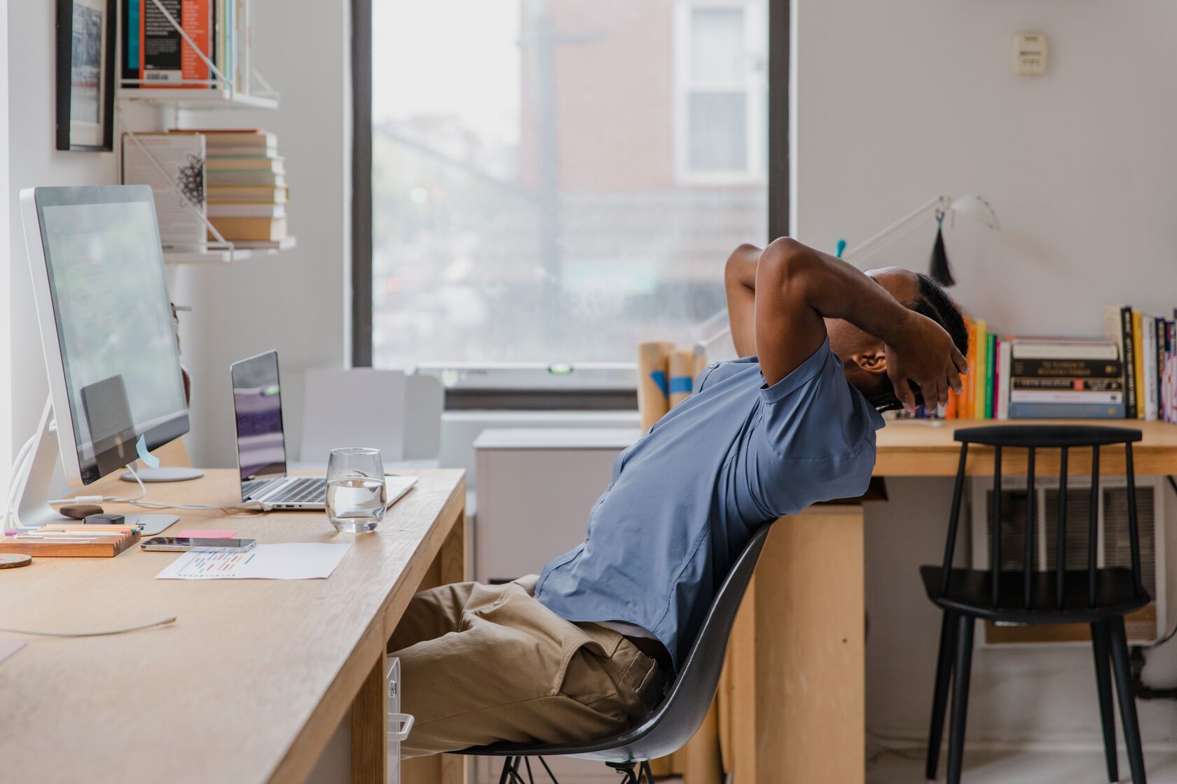 Man in a blue shirt leaning back in a chair at a bright office desk with his hands behind his head