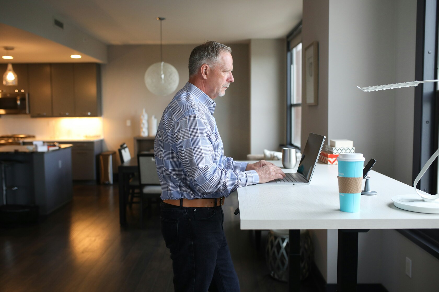 Man working at a laptop on a standing desk beside a tall window in a bright apartment
