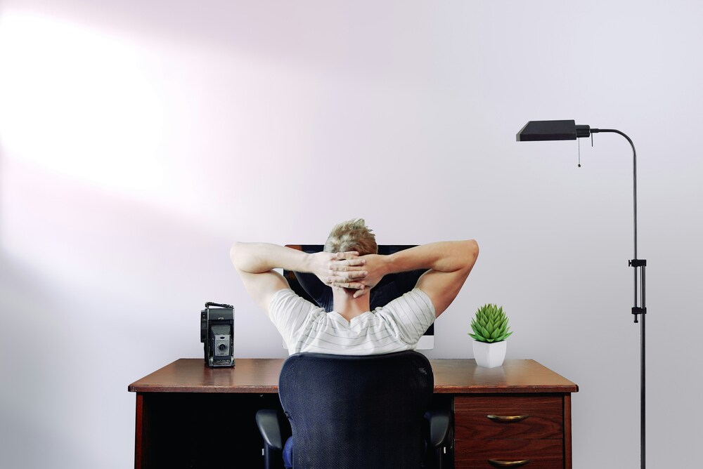 Person seen from behind sitting at a wooden desk with hands behind the head next to a floor lamp and a small plant